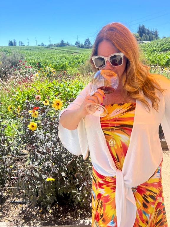 Gina Birch in a yellow dress sipping wine in a vineyard with flowers