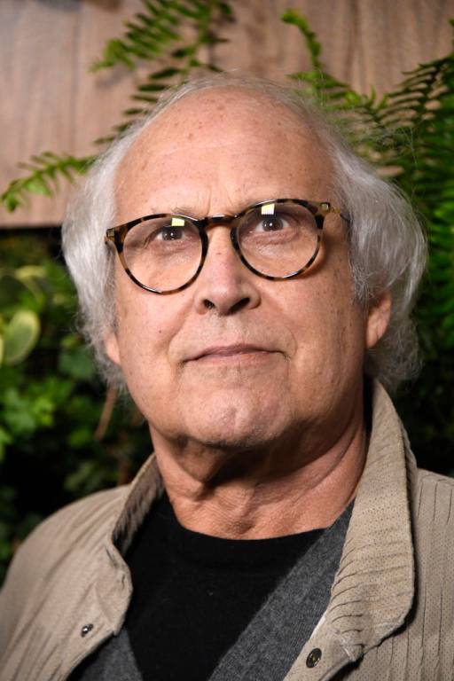 Chevy Chase attends the Global Green 2019 Pre-Oscar Gala with white hair on the sides wearing black thick-framed glasses and a khaki cardigan.