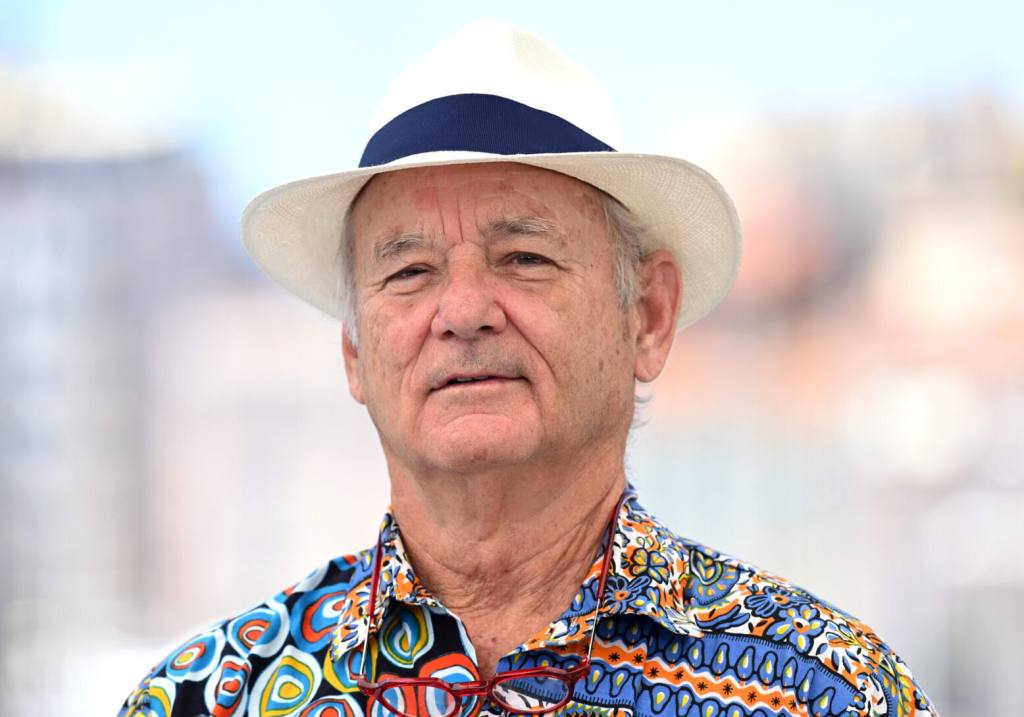 Bill Murray attends the "The French Dispatch" photocall wearing a cream fedora with blue trim peering to the left with a wild patterned shirt on.