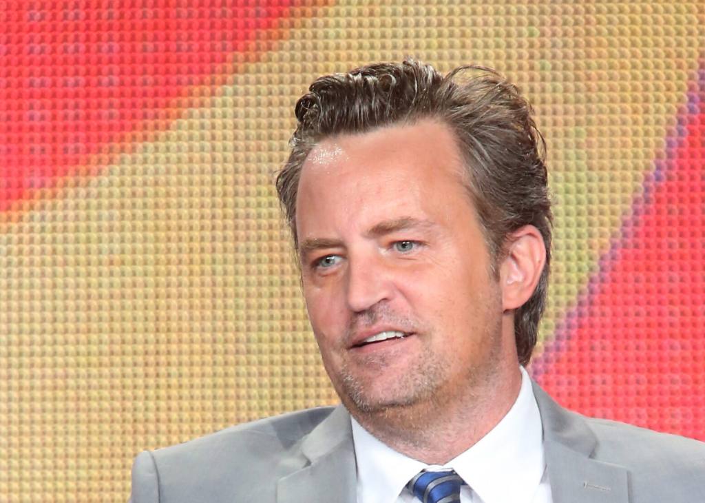 Matthew Perry speaks onstage during 'The Odd Couple' panel at 2015 Winter TCA Tour - Day 6 facing left wearing a grey suit.