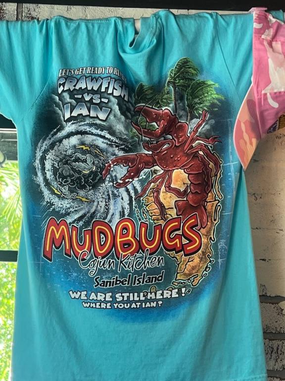 Blue MudBugs t-shirt inscribed with "we are still here" and a drawing of a hurricane