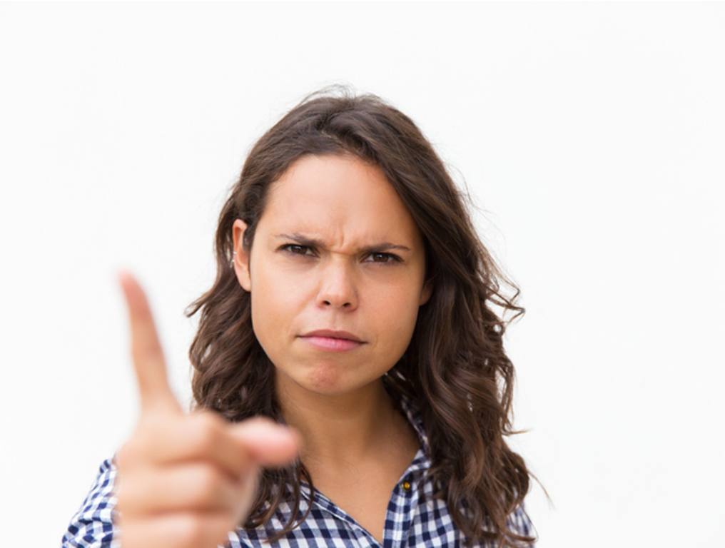 Strict frowning Latin woman shaking finger at camera. Young woman in casual checked shirt standing isolated over white background, following rules concept.