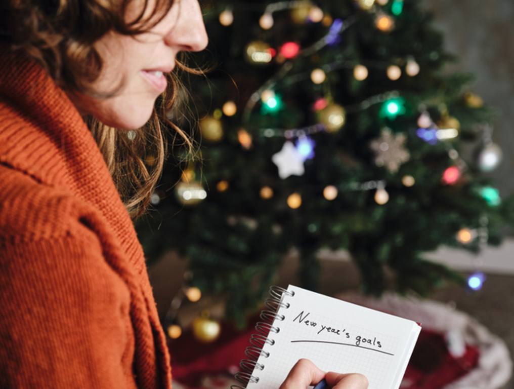 Caucasian woman smiling with orange sweater in profile with new year's goals notebook in hand with unfocused christmas tree in the background