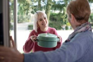 woman opening her front door to another woman holding a casserole dish and smiling with Random Acts Of Kindness