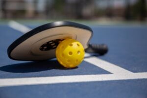 pickleball paddle with a yellow pickleball underneath it on a grey court.