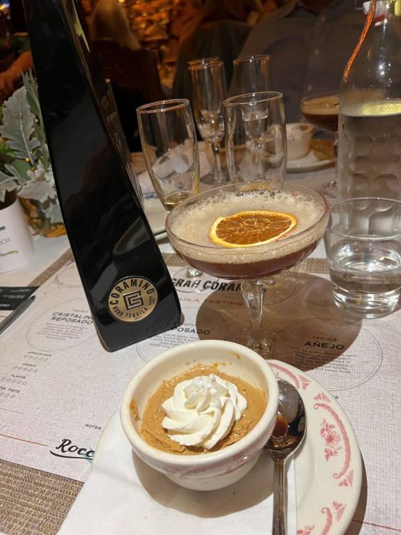 Bottle of Anejo Tequila, cocktail with a dried orange floating on the top and a small ramakin of tan pudding topped with white shipped cream.