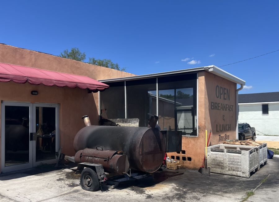 Look at the outside of a BBQ restaurant with a screened in porch and larger smoker.