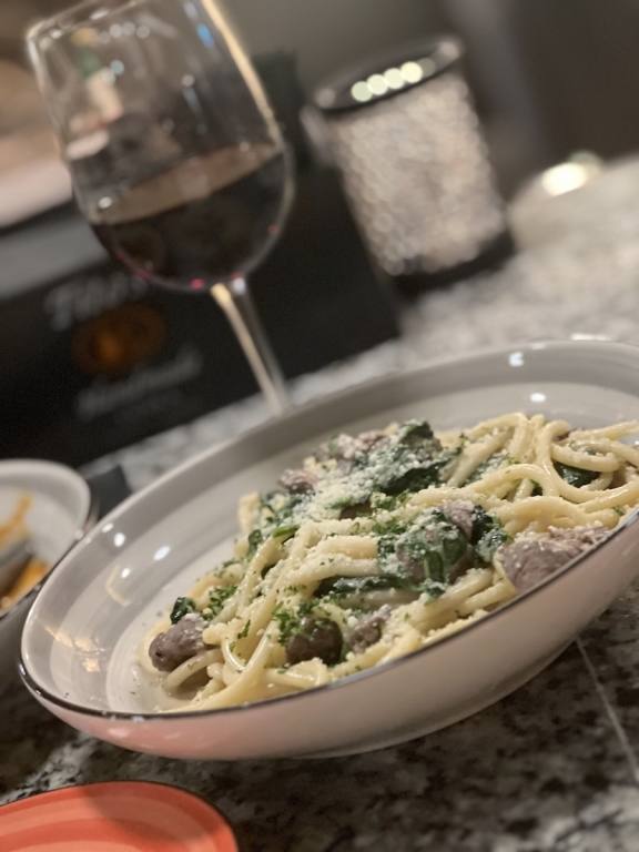 Bowl of thick noodle pasta with small pieces of meat and some spinach mixed in, a glass of red wine in the background