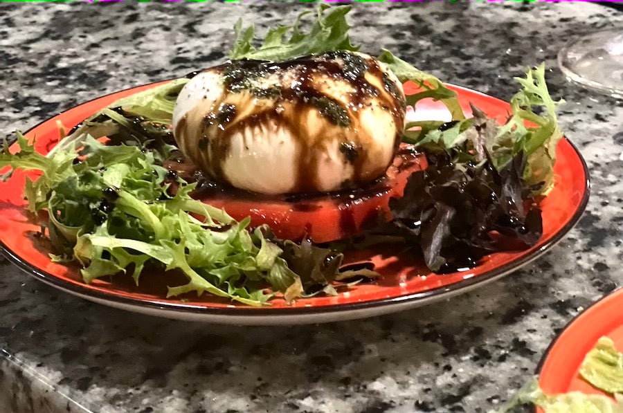 Photo of burrata cheese on a red plate drizzled in dark balsamic vinegar and surrounded by greens for a Sizzle Dining Stop at La Fontanella