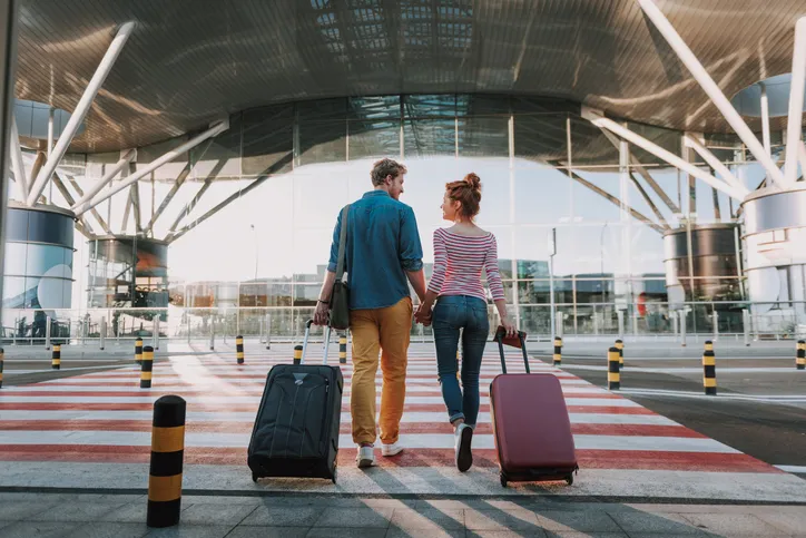 Full length back view portrait of young man and his charming girlfriend walking and carrying their trolley bags. They looking at each other and smiling, time to book Thanksgiving travel from RSW