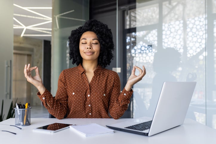 African American woman in a brown polka dot shirt meditating with eyes closed in front of a laptop at her modern office space, promoting wellness and mindfulness at workplace.