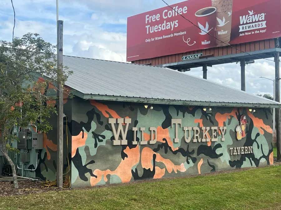 Outside of a bui8lding painted in camouflage and letters that read Wild Turkey Tavern