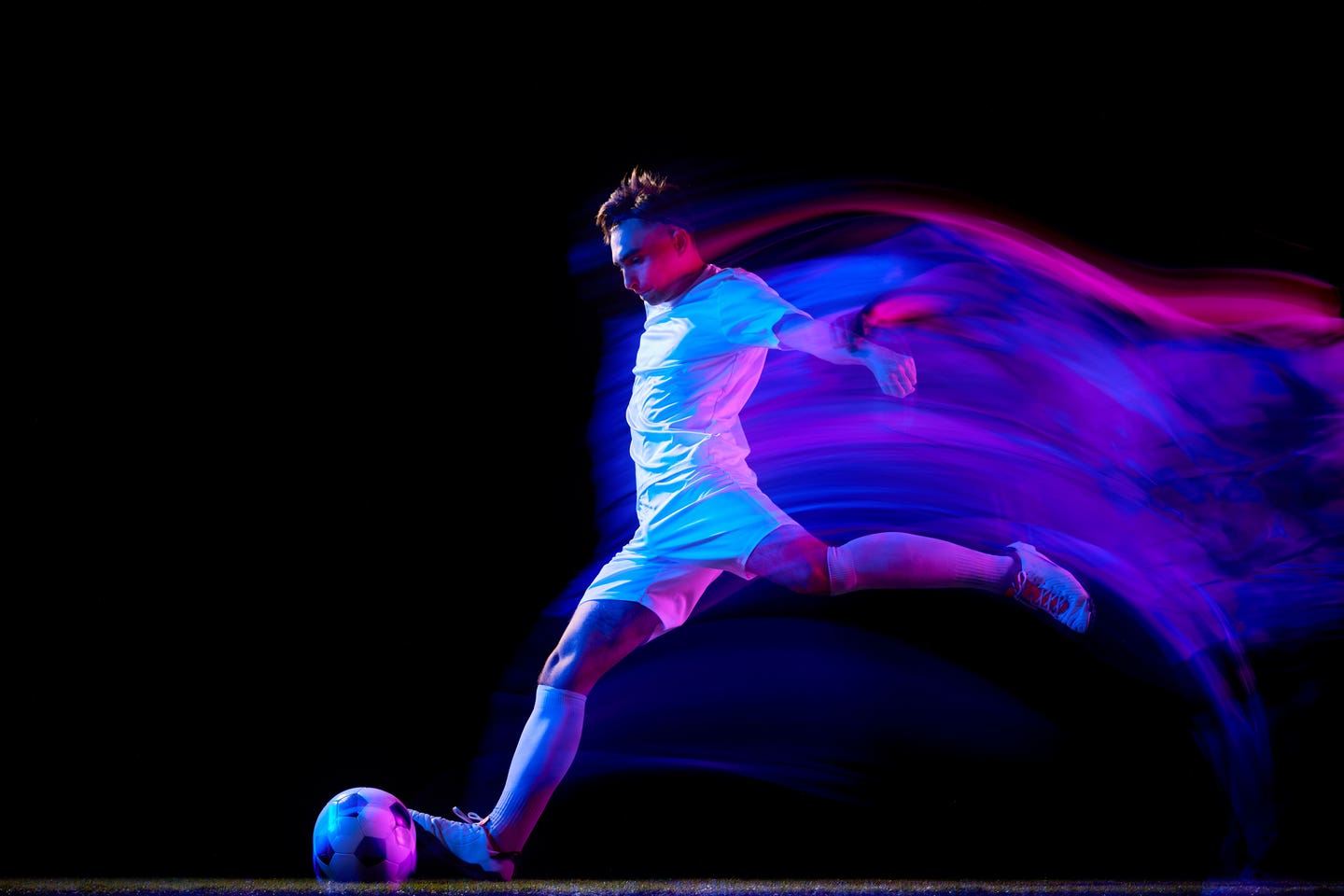 Focused football player, dressed in white under neon lighting, performs swift mid-air kick, mastering agility and technique in his practice. Motion blur. Concept of championship, sport events. Ad