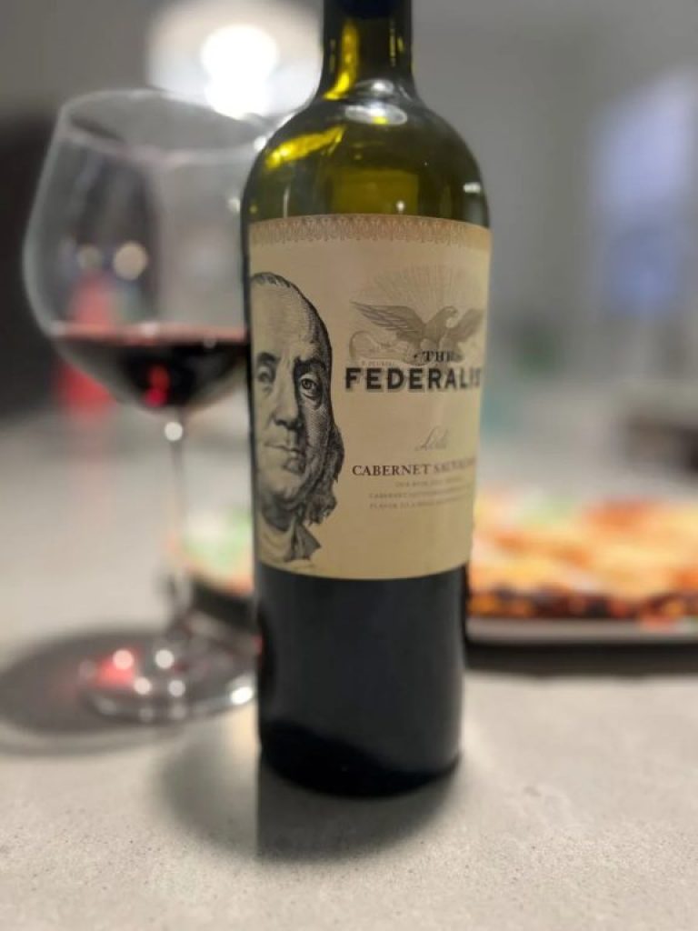 A bottle of The Federalist wine, a wine glass with red wine in it, and a blurred pizza in the background. good sips for National Red Wine Day and Labor Day